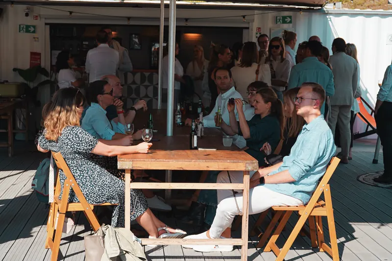 Gäste an einer langen Holztafel in der Abendsonne vor der Bar